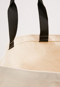 Torba tote z płótna w naturalnym beżowym kolorze z czarnymi materiałowymi paskami. Wnętrze jest gładkie i niepodszewkowane, z widocznymi detalami szwu.