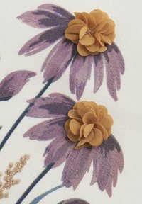 Blumiges Textildesign mit lila Blumen und geschichteten gelben Stoffakzenten. Cremerfarbenen Hintergrund mit subtilen glitzernden Elementen.