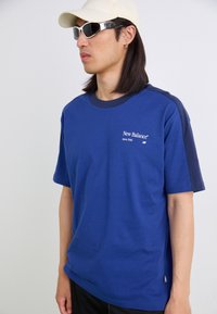 T-shirt en coton bleu avec des manches contrastantes bleu marine, arborant le logo blanc "New Balance". Le modèle porte des lunettes de soleil et une casquette de couleur claire.
