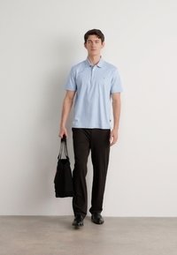 Camisa polo azul-claro de mangas curtas, combinada com calças pretas. A pessoa segura uma mala tote preta e está a usar sapatos pretos.