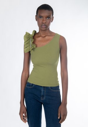 Jonge vrouw die een asymmetrische top met ruches in olijfgroen draagt, versierd met kristallen, en een donkerblauwe spijkerbroek, staand tegen een witte achtergrond.