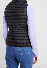 Kobieta ubrana w czarną pikowaną kamizelkę bez rękawów na lawendowej koszuli z długim rękawem i niebieskich dżinsach, widziana od tyłu.