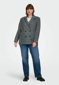 Femme portant un blazer croisé à carreaux gris, un haut blanc, un jean bleu et des chaussures noires, se tenant devant un fond clair.