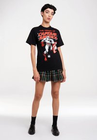 Schwarzes T-Shirt mit dem Grafik "Die Herkunft von Captain America" in Rot und Weiß. Kombiniert mit einem grünen karierten Faltenrock. Schwarze Schuhe und Socken.