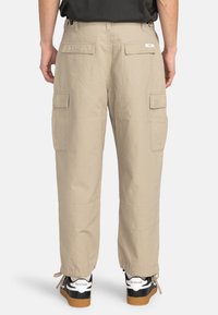 Beige cargo bukser lavet af letvægtsstof, med flere sidelommer og elastiske manchetter, parret med sorte sneakers.