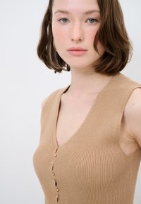 Beige geripptes Tanktop mit tiefem V-Ausschnitt und fünf kleinen goldenen Knöpfen auf der Vorderseite. Die taillierte Passform und der ärmellose Stil betonen die Schultern.