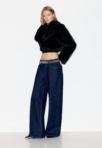 Frau steht vor einem schlichten weißen Hintergrund, trägt eine schwarze, kurz geschnittene Jacke aus Kunstpelz, dunkle Jeans mit weitem Bein und dekorativem Gürtel sowie spitze schwarze Schuhe.