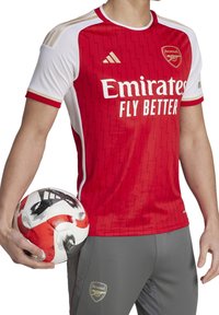 Red Arsenal jersey with white accents, featuring a zigzag pattern, Adidas logo, and "Emirates Fly Better" text. Gray pants and soccer ball.