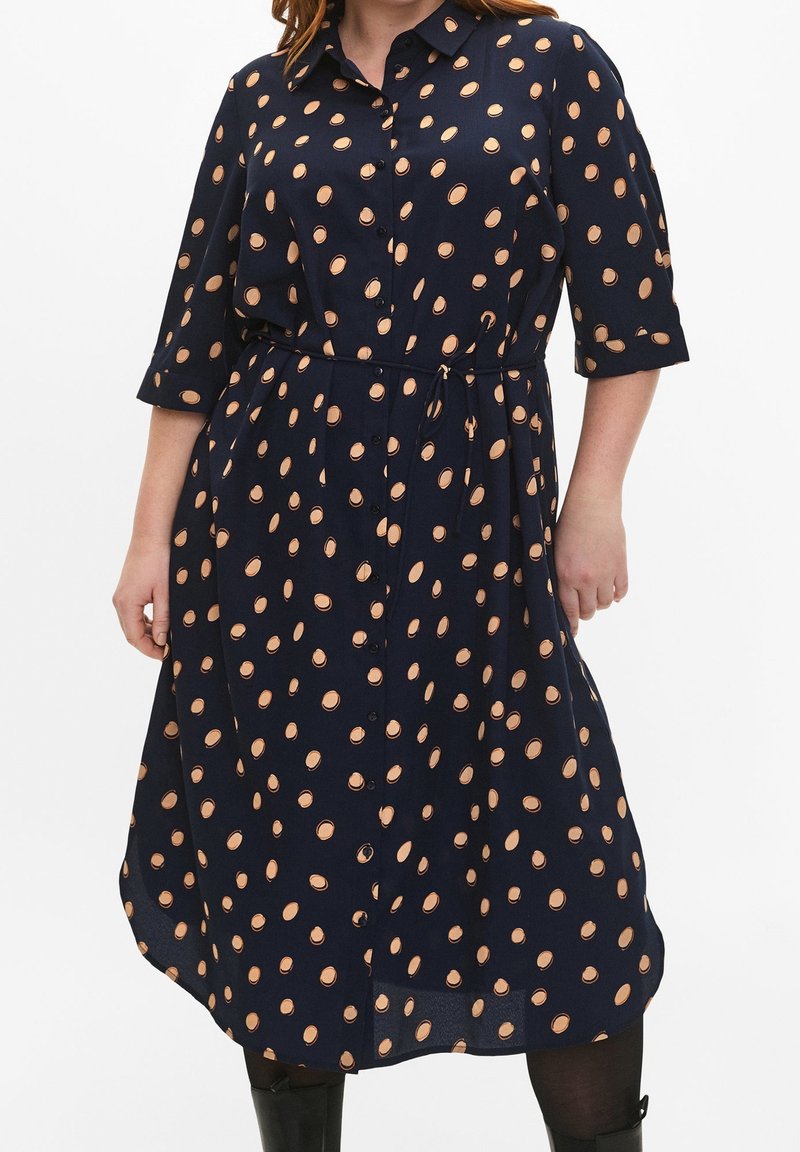 Robe bleu marine longueur genou avec des pois pêche, boutonnée devant, manches trois-quarts et ceinture nouée à la taille, portée par une personne.