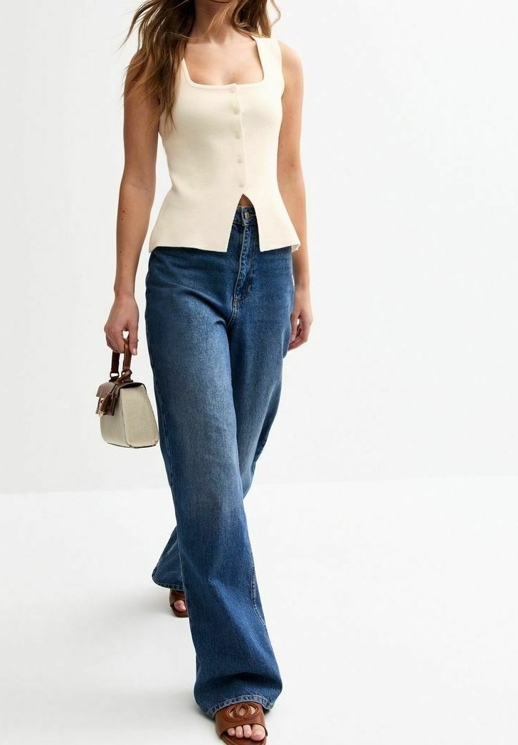 Kobieta ubrana w bezrękawnikowy kremowy top na guziki, szerokie niebieskie dżinsy oraz brązowe sandały, trzymająca małą beżową torebkę z brązowymi uchwytami.