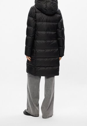Personne portant un manteau matelassé noir à capuche et un pantalon gris à jambes larges, debout de dos devant un fond blanc uni.