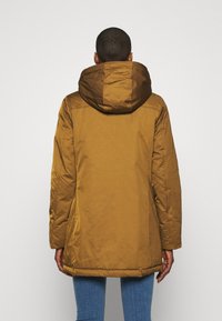 Persoon met kort haar, gekleed in een mosterdgele hoodie en blauwe spijkerbroek, staand met de rug naar de camera tegen een effen achtergrond.