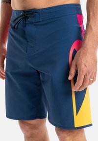 Niebieskie szorty kąpielowe z żółtym i różowym wzorem graficznym, wykonane z gładkiego materiału, z zapięciem na sznurek i boczną kieszenią.