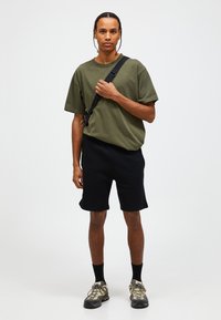 T-shirt in cotone verde oliva, pantaloni corti neri e calzature beige. La T-shirt ha una vestibilità comoda; i pantaloni corti sono lunghi fino al ginocchio con risvolti semplici.