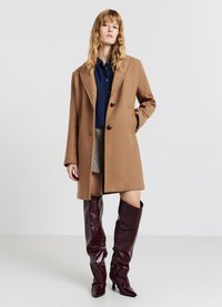 Cappotto in lana marrone, con texture, colletto a punte, due bottoni e tasche laterali. Abbinato a una camicia di jeans scura e stivali alti lucidi color bordeaux.