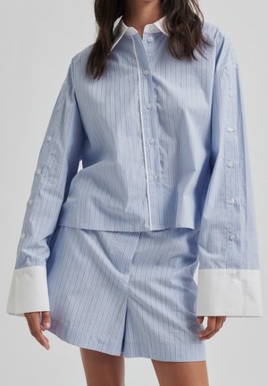 Vrouw draagt een lichtblauw gestreept overhemd met knopen, met oversized witte manchetten en bijpassende korte broek, tegen een effen grijze achtergrond.