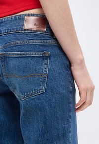 Lähivõte inimesest, kes kannab siniseid Pepe Jeansi teksaseid, millel on nähtav tagatasku ja pruun nahkbrändi silt, riietatud punasesse toppi.