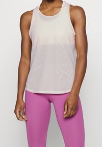 Kobieta w jasnoróżowej, bezrękawnikowej bluzce z siateczki i jasnoróżowych legginsach, stojąca na jednolitym białym tle.