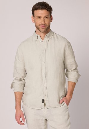 Mężczyzna o krótkich kręconych włosach, ubrany w jasnobeżową koszulę na guziki z podwiniętymi rękawami oraz dopasowane spodnie, stojący z jedną ręką w kieszeni.