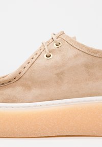 Chaussure en daim beige à bout arrondi, dotée de deux œillets dorés, de coutures crème et d'une semelle texturée orange clair.
