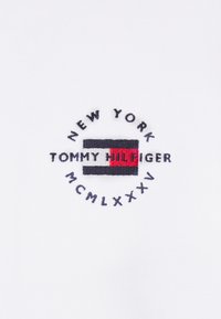Un modello in cotone bianco presenta un logo circolare ricamato con la scritta "NEW YORK," "TOMMY HILFIGER," e "MCMLXXXV" nei colori blu navy e rosso.