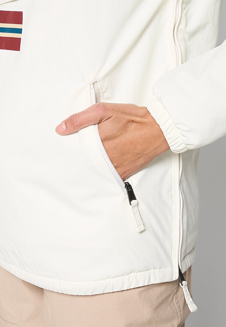 Hand in der Reißverschlusstasche einer weißen Jacke mit elastischen Bündchen, dabei sind beige Hosen sichtbar.