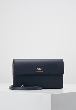 Pochette Emporio Armani bleu marine avec rabat, logo doré et bandoulière amovible, posée sur un socle blanc devant un fond gris clair.
