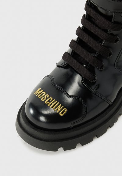 Stivale in vernice nera con lacci neri e suola spessa nera. Presenta il marchio dorato "MOSCHINO" sulla punta. Finitura lucida e strutturata.