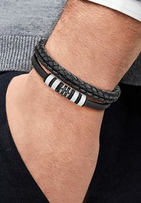 Bracciale in pelle con fili neri intrecciati e dettagli in argento. Presenta un centro testurizzato e un design elegante e moderno.