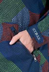 Ruka částečně v kapse barevné vzorované bundy s modrými, zelenými, červenými a světle modrými motivy a s vyšitým nápisem "HYRAR" na manžetě.