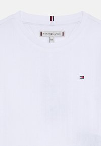 T-shirt blanc à côtes avec col rond, arborant un petit logo multicolore sur le côté gauche de la poitrine. Tissu doux avec des détails de texture subtils.