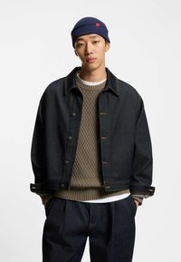 Dunkelblaue Jeansjacke mit Kragen, Frontknöpfen und Taschen, über einem strukturierten braunen Pullover getragen. Eine blau gerippte Strickmütze vervollständigt das Outfit.
