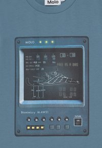Blå t-shirt med ett grafik av en kontrollpanel med mätare, etiketter och en skissad flygplan. Texten lyder "FRI SOM EN FÅGEL."