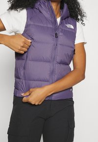 Fioletowa pikowana kamizelka z wysokim kołnierzem, kieszeniami na zamek i zapięciem na zamek błyskawiczny. Lekki materiał, pikowany wzór, odpowiednia do noszenia w warstwach.