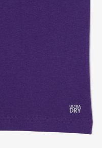 Camiseta de algodón morada con una textura suave, que presenta un dobladillo cosido y un pequeño logo "ULTRA DRY" en blanco en la esquina inferior derecha.