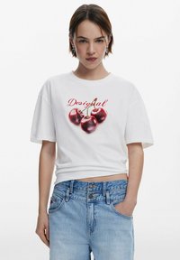 White cotton t-shirt with loose fit, featuring a graphic of three red cherries and the word "Desigual" in red text. Paired with light blue jeans.
