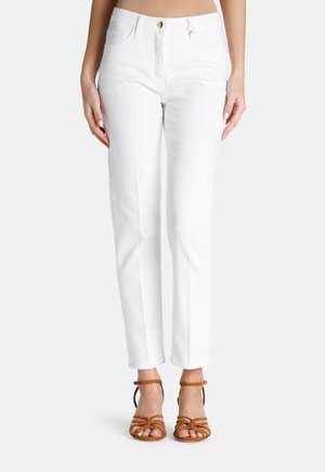 Pantalones hasta el tobillo de color blanco con un diseño ajustado, que cuentan con bolsillos frontales y un cierre de botón dorado. Combinados con sandalias marrones de tiras.