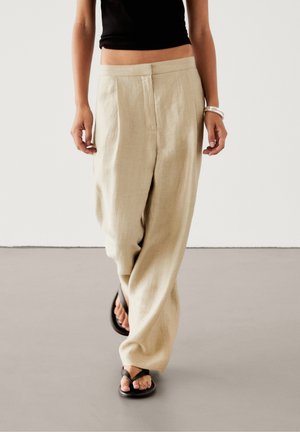 Beige linnedragter med løs pasform, der har forreste folder, høj talje og glat tekstur, kombineret med sorte klipklapper.