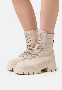 Botas de encaje beige con parte superior texturizada, cuello acolchado y suela gruesa y ranurada con ojales metálicos y detalle de tirador.