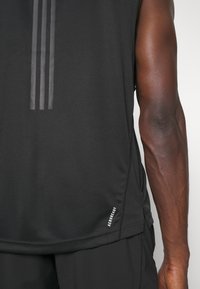 Gros plan d'une personne portant un maillot de sport noir sans manches avec trois bandes verticales et le texte « AEROREADY » sur la partie inférieure.