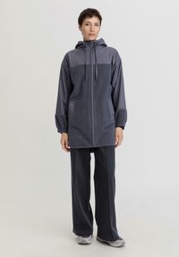 Giacca isolante grigia con cappuccio, dotata di un pannello superiore in contrasto, chiusura frontale con zip, polsini elasticizzati e tasche laterali. Abbinata a pantaloni grigio scuro.