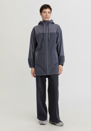 Grijze geïsoleerde jas met een capuchon, voorzien van een contrasterend bovenpaneel, een ritssluiting aan de voorkant, elastische manchetten en zijzakken. Gepaard met donkergrijze broeken.