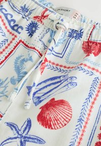 Gros plan sur un short blanc avec des imprimés marins bleus et rouges comprenant des hippocampes, des poissons, des étoiles de mer, des coquillages et une taille élastiquée avec cordon.