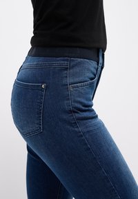 Dunkelblaue Denim-Jeans mit einem mittelhohen Bund, schmaler Schnitt und zwei Gesäßtaschen mit silbernen Druckknöpfen. Glatte Textur, minimale Verblassung.