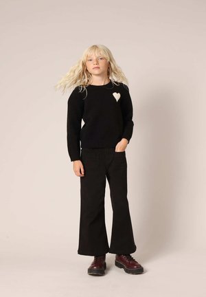 Zwarte trui met een crèmekleurig hartdetail, geribde halslijn en onderrand. Gecombineerd met zwarte wijde broek en bordeauxrode veterschoenen.