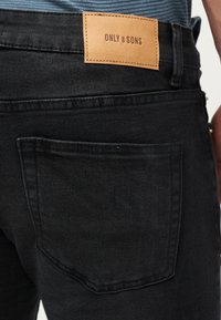 Οπίσθια όψη μαύρου τζιν με κεντημένη ταμπέλα από ταμπά δέρμα με την επιγραφή «ONLY & SONS» πάνω από την δεξιά πίσω τσέπη.