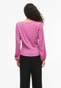 Kvinne med langt, kr�øllete hår som har på seg en teksturert, rosa langermet topp og løse, svarte bukser, sett bakfra mot en hvit bakgrunn.