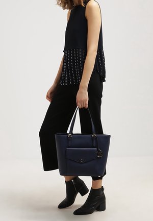 Marineblauwe draagtas met een voorvak en gouden beslag, gecombineerd met een zwarte mouwloze top en wijde broekspijpen, aangevuld met enkellaarzen.