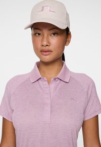 Giovane donna con capelli lisci e scuri indossa un cappellino da baseball rosa chiaro e una polo abbinata con colletto, il tutto su uno sfondo bianco puro.