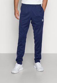 Tmavě modré sportovní kalhoty vyrobené z lehké tkaniny, s bílými bočními pruhy a elastickým pasem. V kombinaci s bílými teniskami.